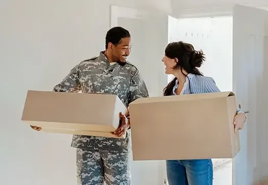 Man in military uniform and woman carrying moving boxes inside a home