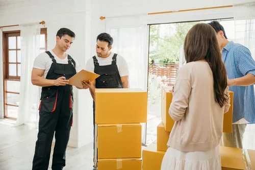 Two movers check a clipboard while helping a family pack and move boxes inside a bright, modern home.